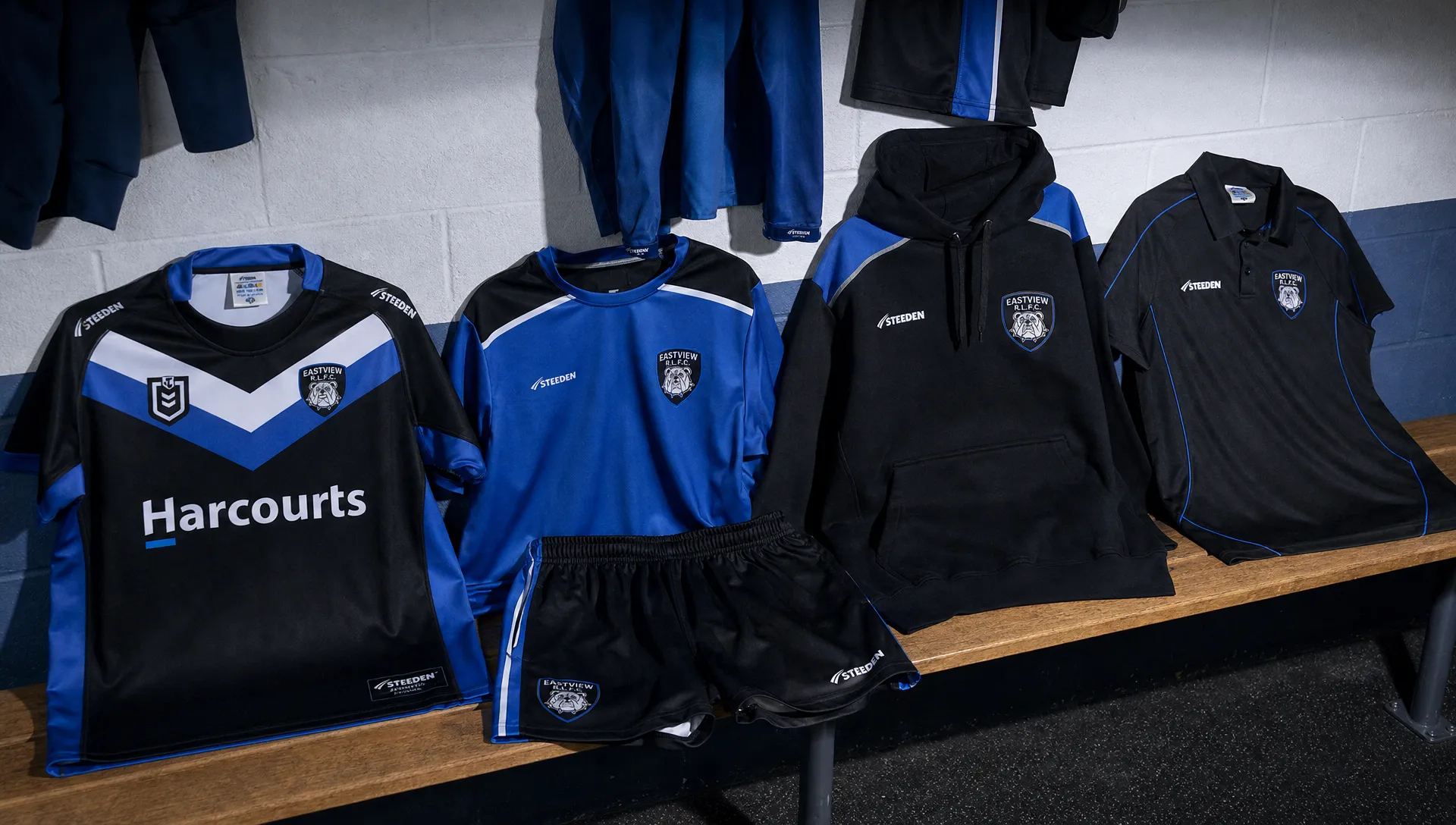 A rugby league club apparel collection laid out on a bench in a changing room, with a jersey, training top, shorts, hoodie and polo ready for players and supporters.