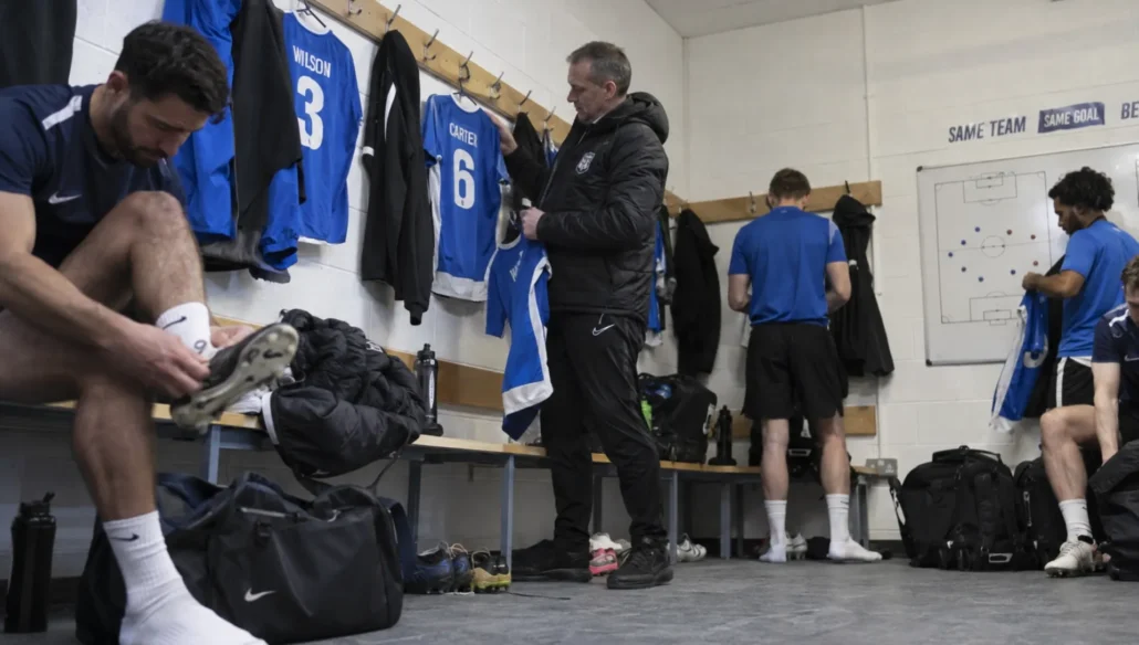 A team manager checks personalized soccer jerseys as players collect their kits in the club equipment room.